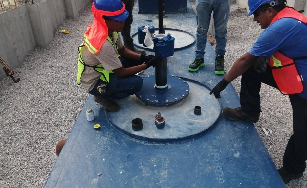 Dos trabajadores con casco azul, chaleco reflejante y guantes montando la válvula y la brida de registro sobre la tapa de una cisterna prefabricada azul.